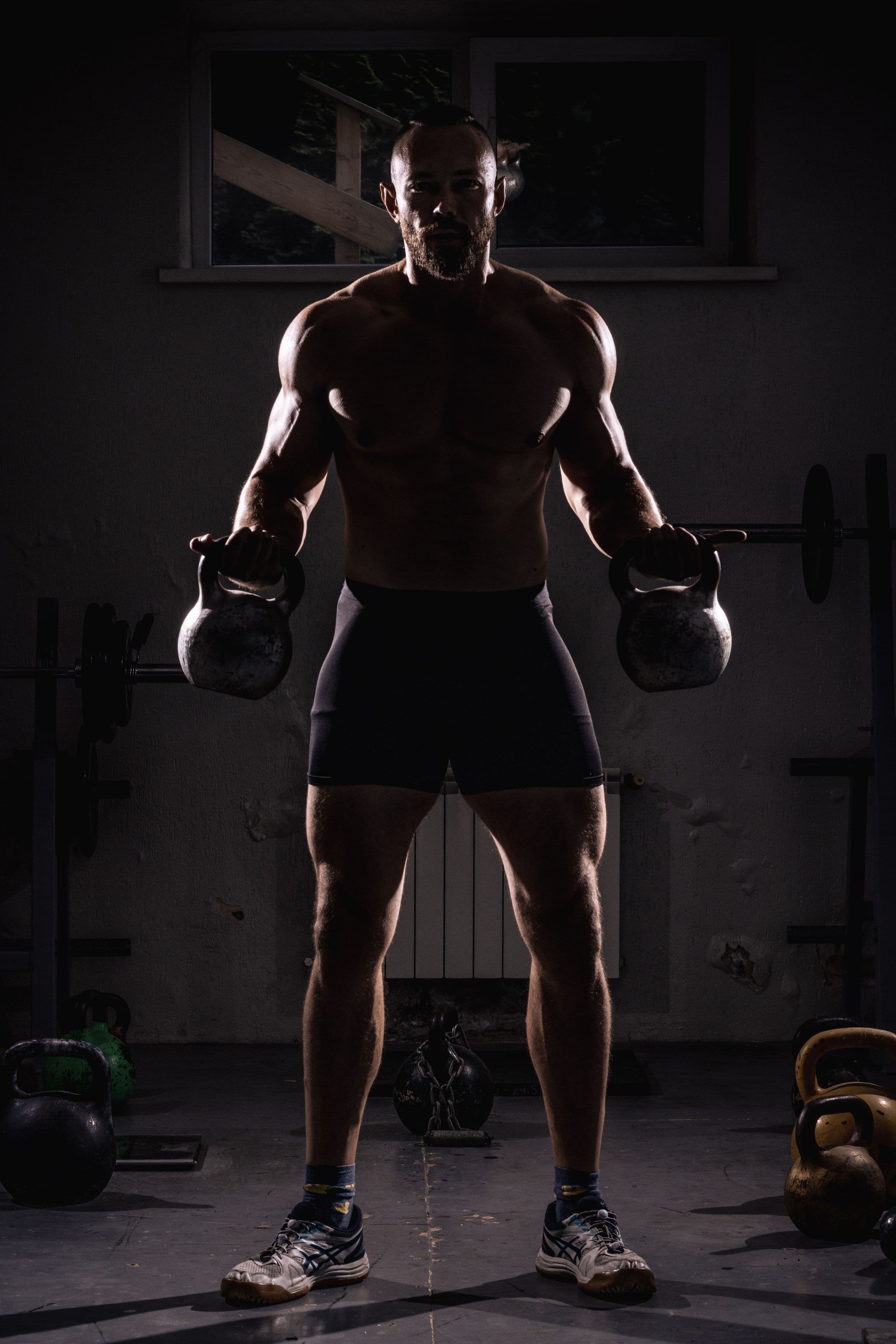 An athlete standing with a kettlebell in each hand under hard side light